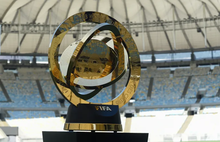 A golden trophy with a hollow, circular design and a central sphere, standing on a black base with the FIFA logo. The trophy is on a field in a large stadium with blue and yellow seats visible in the background.