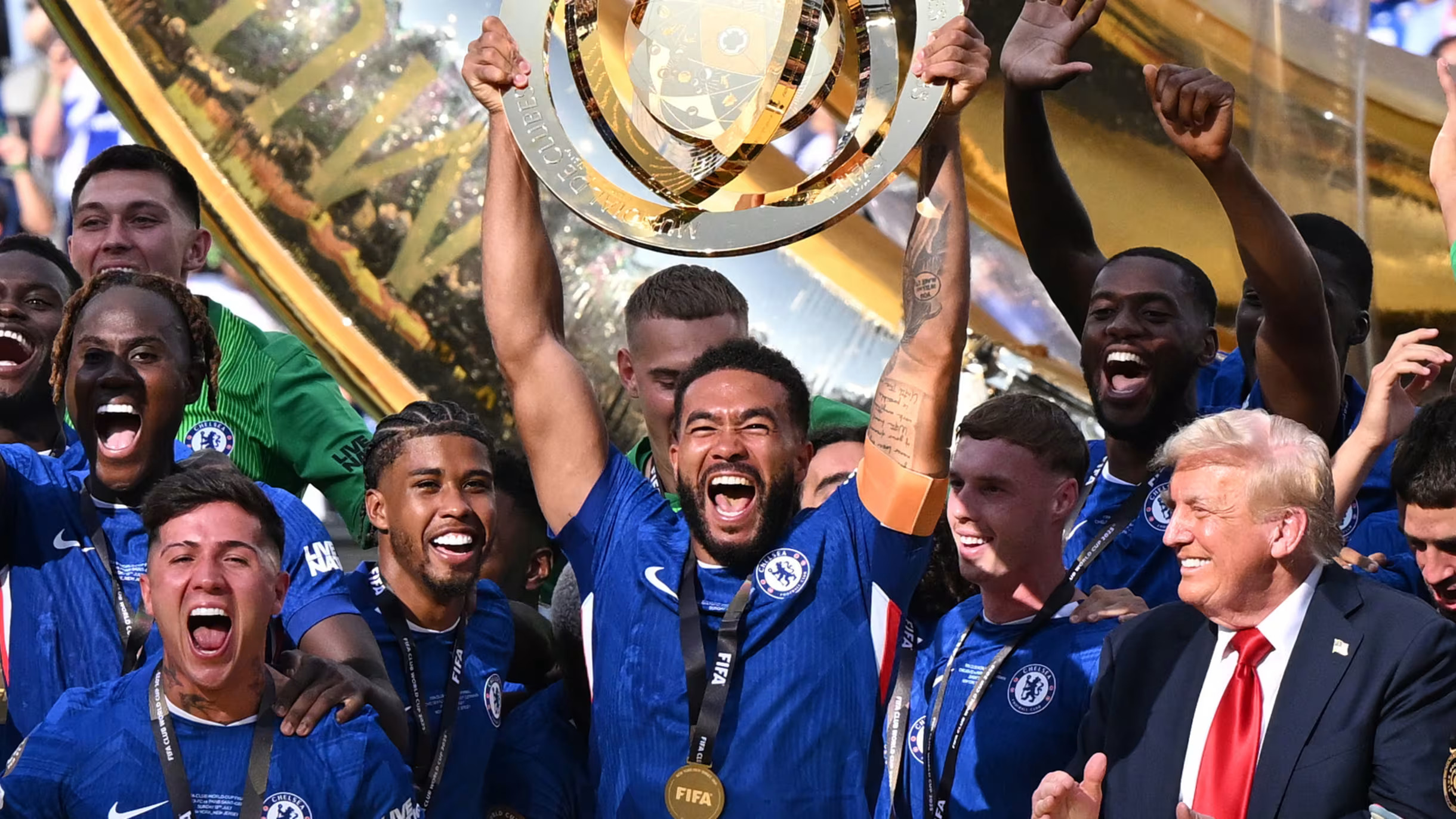 Chelsea players celebrate with the FIFA Club World Cup trophy, cheering and holding the gold shield aloft while surrounded by teammates and staff in blue jerseys.