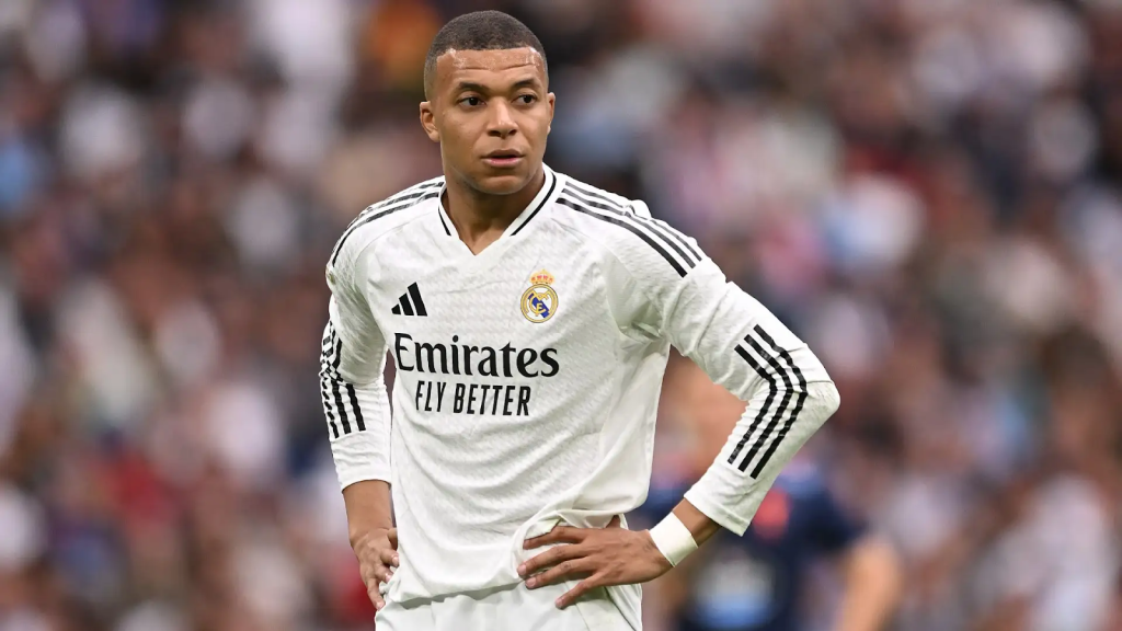 A football player, Kylian Mbappe, wearing a Real Madrid jersey with the logo “Emirates Fly Better” stands on the field with his hands on his hips during a match, with a blurred crowd visible in the background.